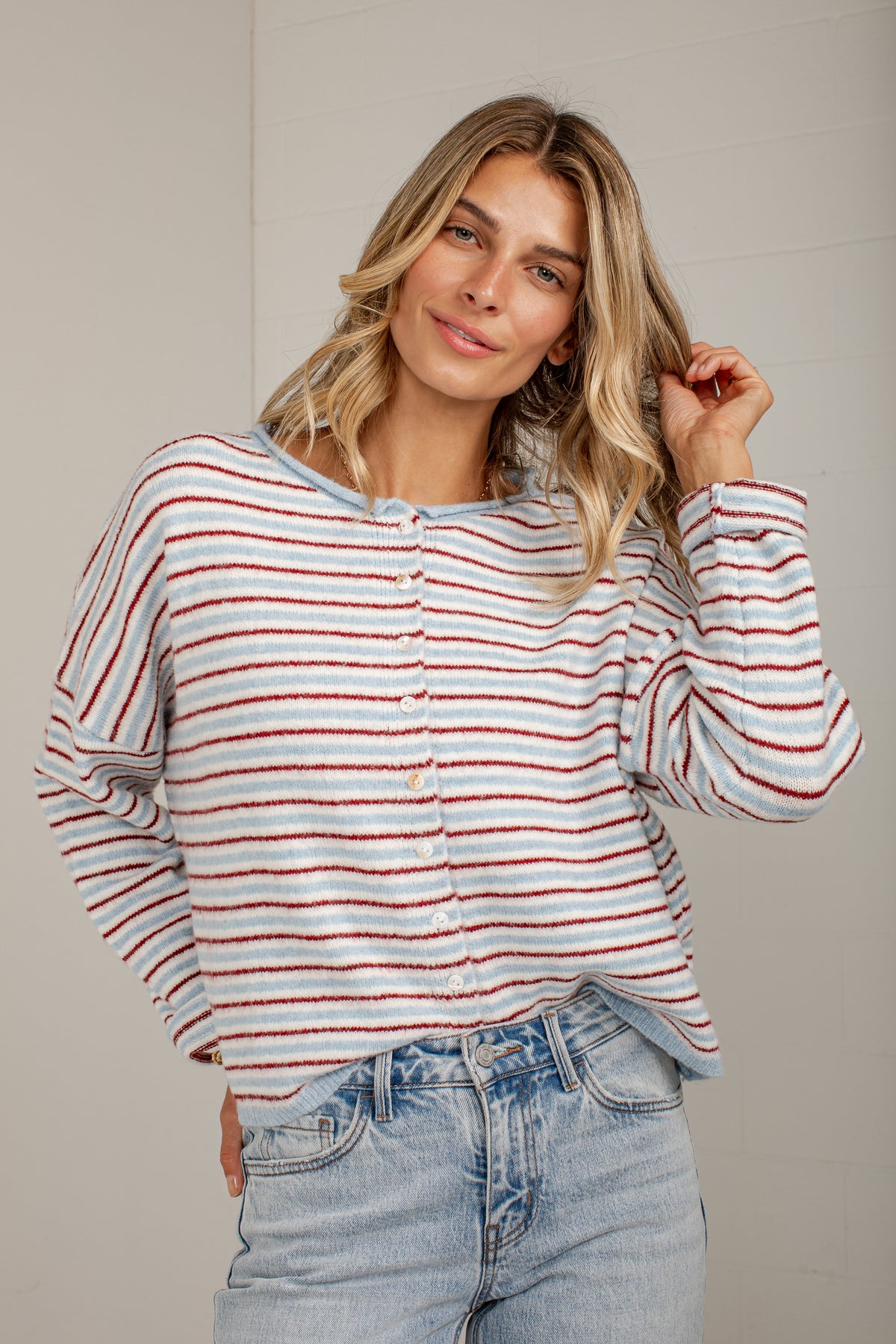 A woman wearing a long-sleeve knit top with thin red and white horizontal stripes, paired with light-wash blue jeans, standing against a neutral light gray wall.