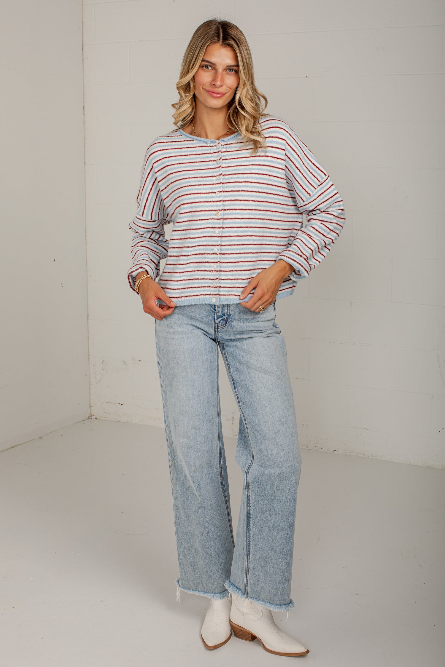 A woman modeling a long-sleeve button-up shirt with thin red and orange horizontal stripes on a white background, paired with light-wash wide-leg jeans and white ankle boots.