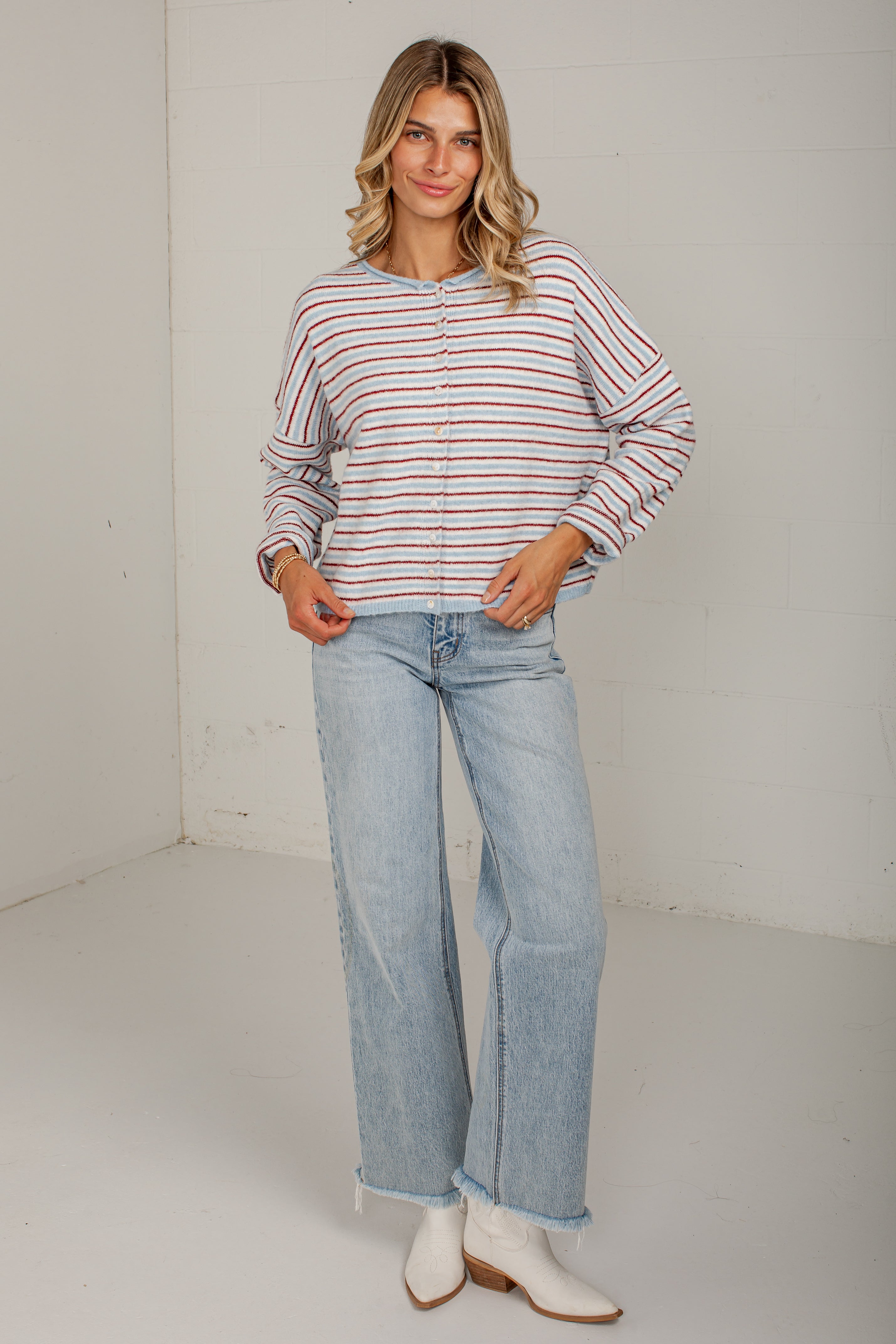 A woman modeling a long-sleeve button-up shirt with thin red and orange horizontal stripes on a white background, paired with light-wash wide-leg jeans and white ankle boots.