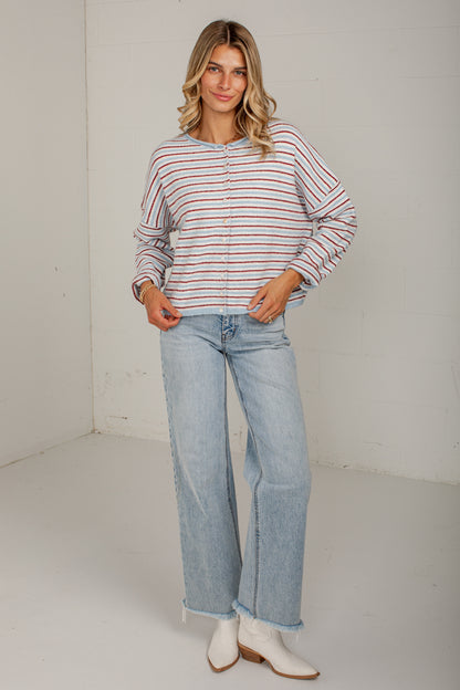 A woman modeling a long-sleeve button-up shirt with thin red and orange horizontal stripes on a white background, paired with light-wash wide-leg jeans and white ankle boots.