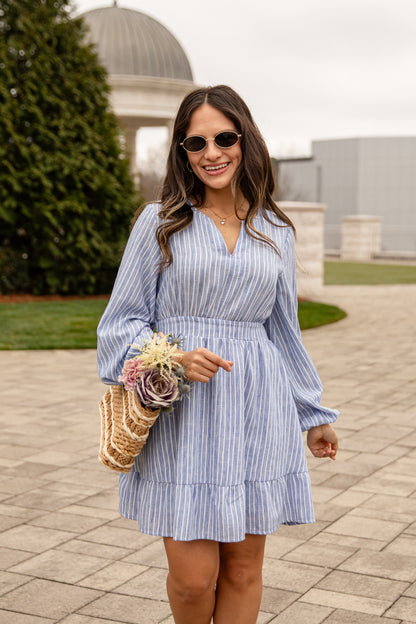 Woman in a blue striped dress holding flowers and a straw bag outdoors.