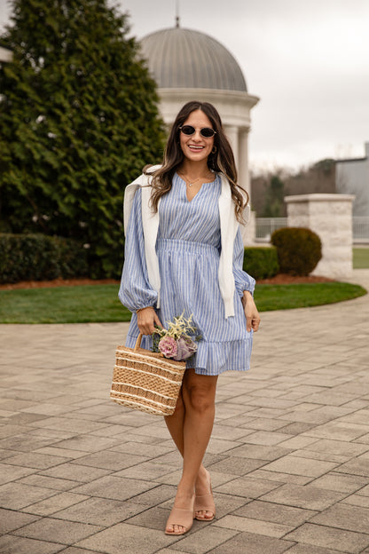 Woman in a blue striped dress with a white cardigan holding a woven bag on a paved walkway.