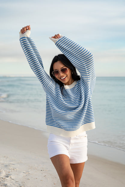 Woman in a blue and white striped sweater and white shorts standing on a beach.