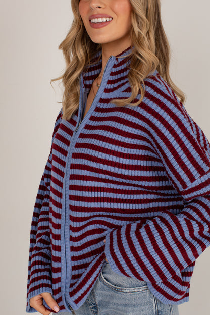 A woman wearing a ribbed knit quarter-zip cardigan with horizontal stripes in dusty blue and burgundy, paired with light-wash blue jeans against a neutral beige background.