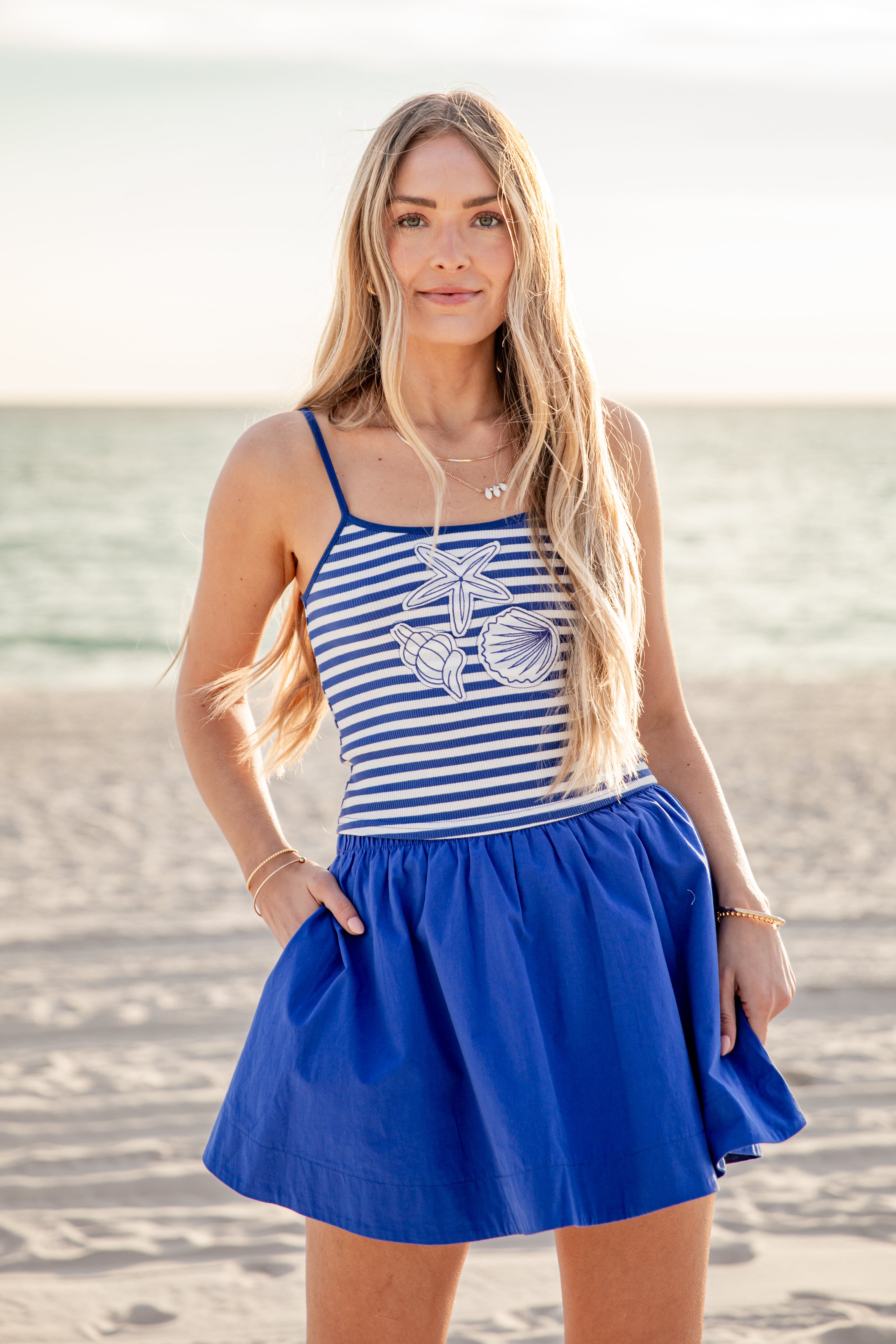 Woman in a striped tank top and blue skirt standing on a beach.