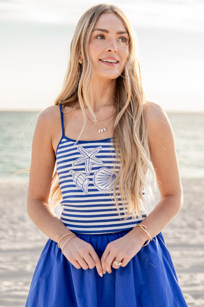 Woman wearing a blue and white striped tank top with shell designs on a beach.
