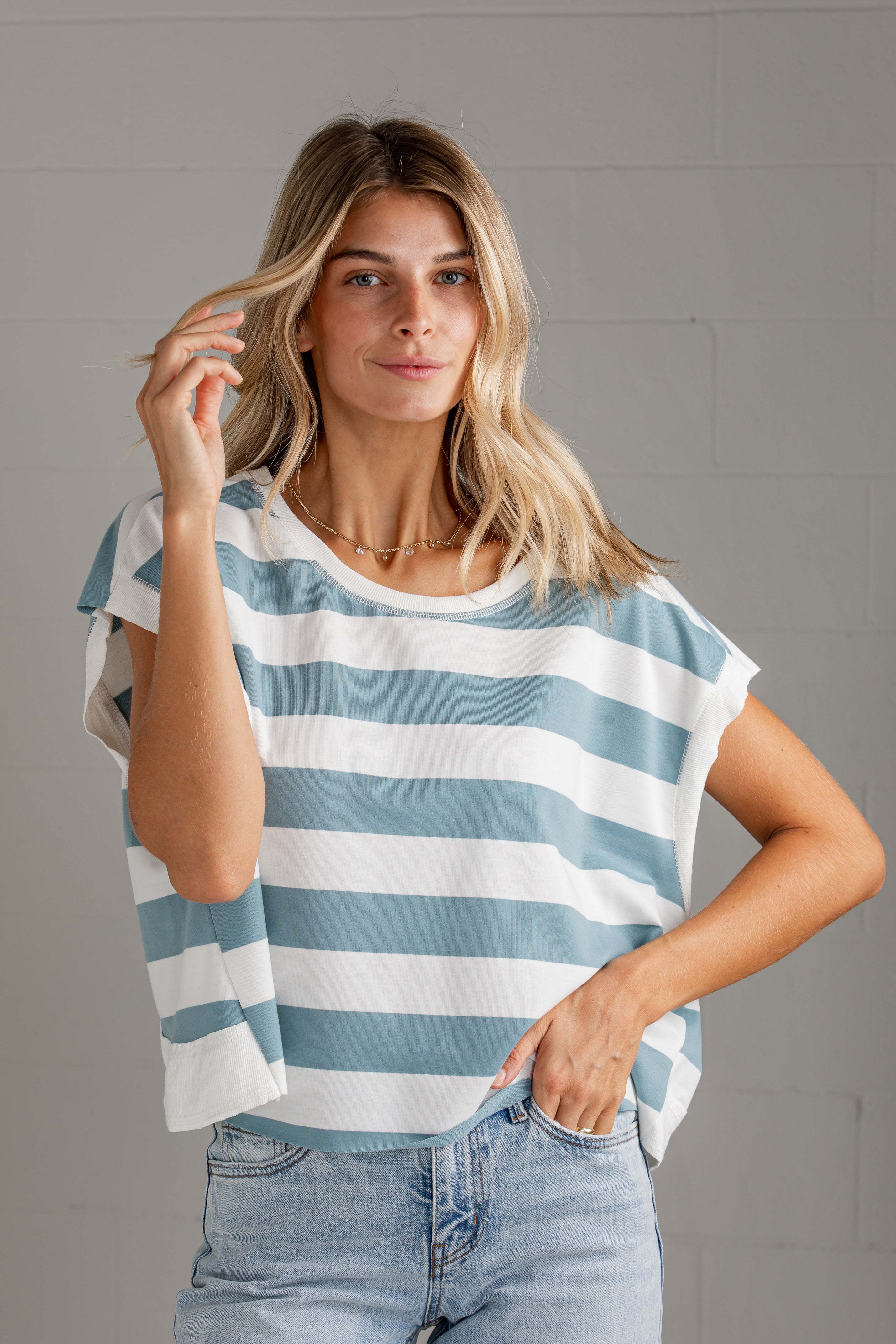 A woman wearing a light blue and white horizontally striped short-sleeve top with a relaxed fit, paired with light-wash blue jeans, standing against a gray brick wall.