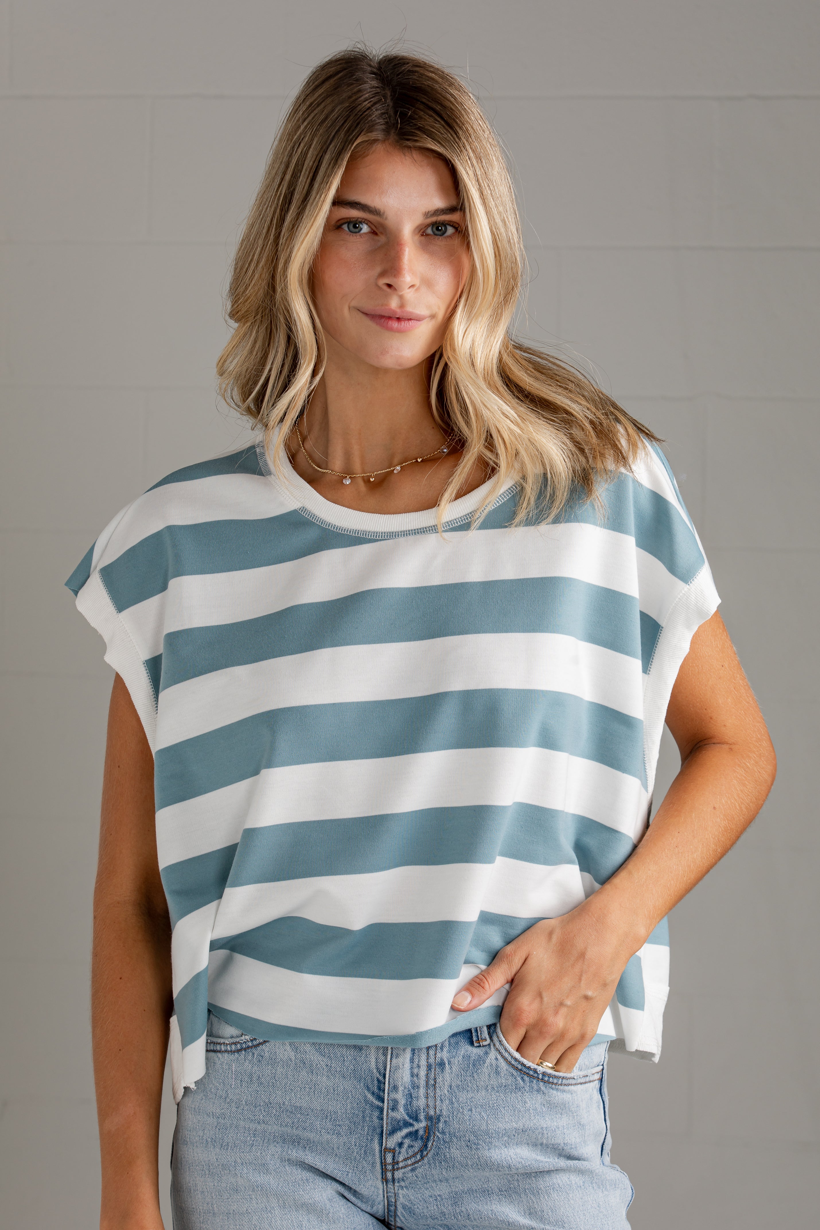 A woman wearing a loose, short-sleeve striped top with horizontal blue-gray and white stripes, paired with light-wash jeans, standing against a light gray wall.