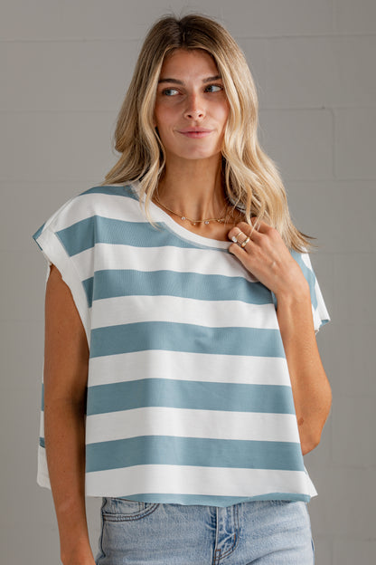A woman wearing a loose, short-sleeve top with horizontal light blue and white stripes, paired with light-wash jeans, standing against a light gray wall.