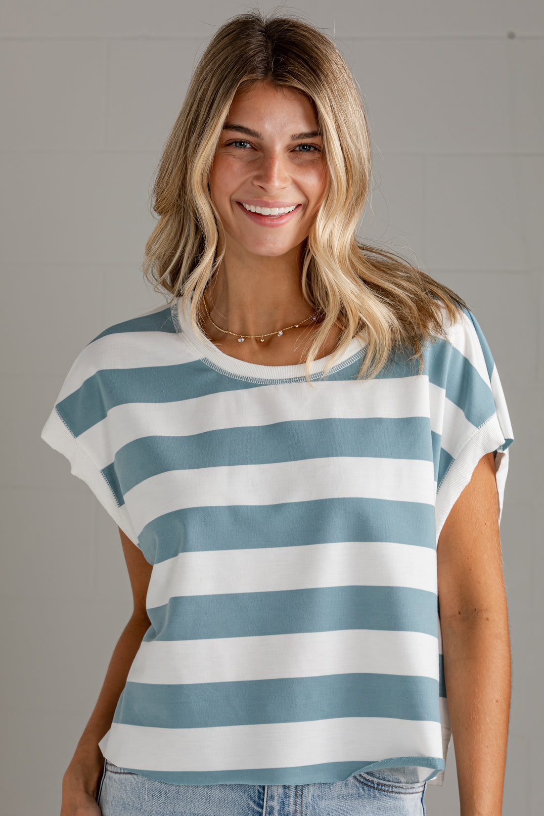 A smiling blonde woman wearing a short-sleeve crewneck top with horizontal light blue and white stripes, paired with light-wash jeans, set against a neutral gray paneled wall.