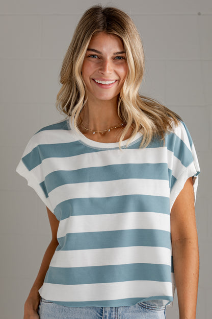 A smiling blonde woman wearing a short-sleeve crewneck top with horizontal light blue and white stripes, paired with light-wash jeans, set against a neutral gray paneled wall.