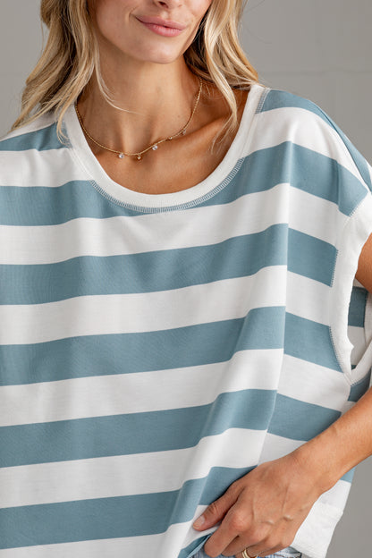 A close-up of a woman wearing a loose, short-sleeve top with horizontal light blue and white stripes. The shot focuses on the chest, neckline, and hand area against a neutral gray background.