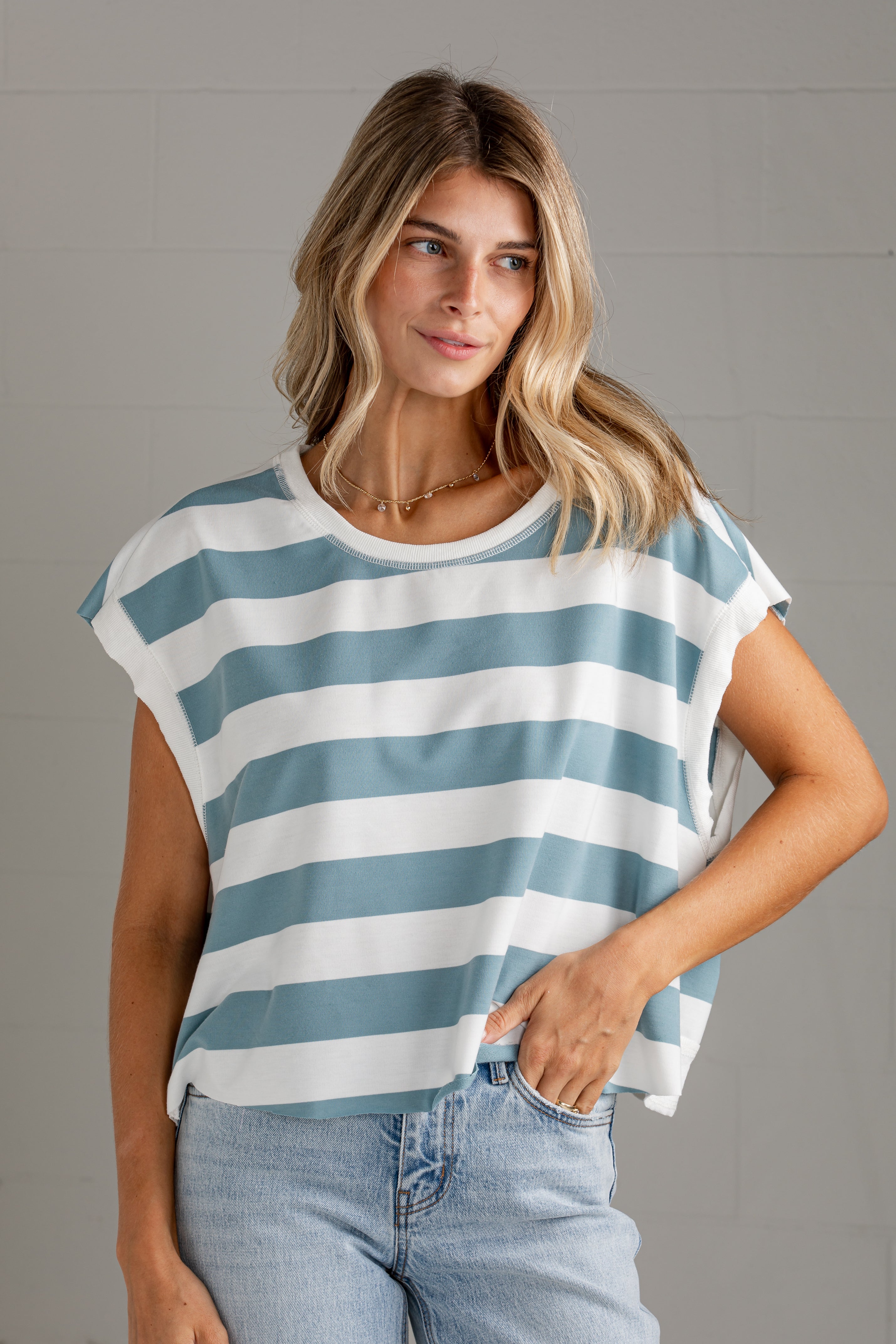 A woman wearing a blue and white horizontally striped short-sleeve crop top with light-wash jeans, standing against a gray brick wall.