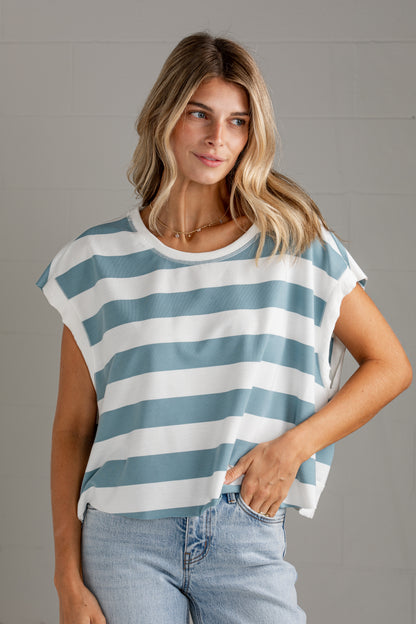 A woman wearing a blue and white horizontally striped short-sleeve crop top with light-wash jeans, standing against a gray brick wall.