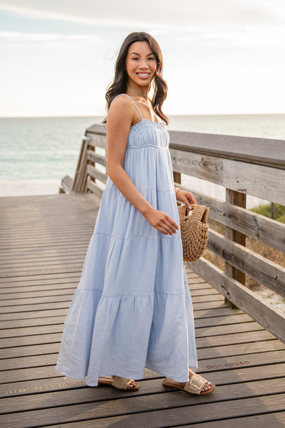 Woman in a light blue dress standing on a wooden pier by the water.