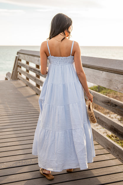 Woman in a light blue dress standing on a wooden boardwalk by the sea.
