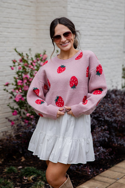 Woman wearing a pink sweater with strawberry patterns and a white skirt outdoors.