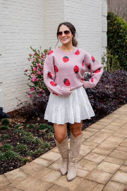 Woman wearing a pink sweater with red fruit patterns, white skirt, and beige boots standing outdoors.