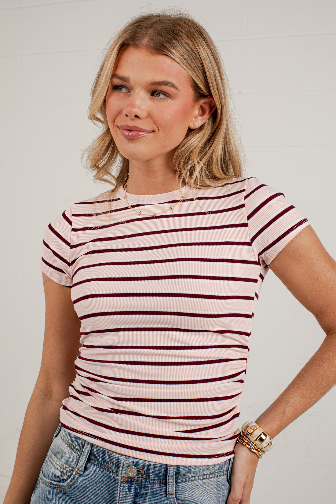 A woman wearing a light pink short-sleeve t-shirt with dark red horizontal stripes, paired with blue jeans, against a white paneled wall.