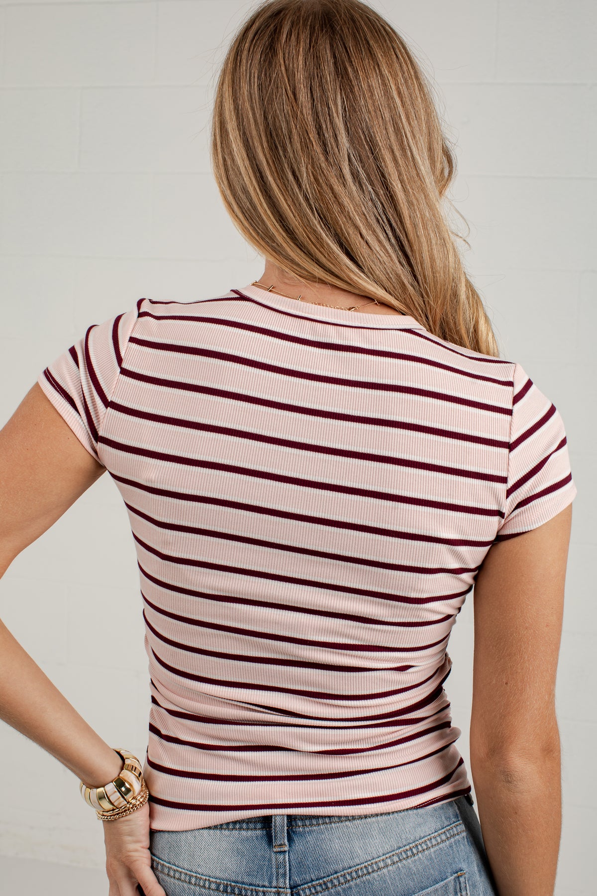 A woman viewed from the back wearing a fitted short-sleeve top with thin horizontal burgundy stripes on a pale pink base, paired with blue jeans against a light gray wall.