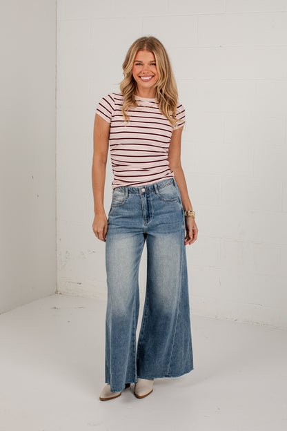 A woman stands in a bright white studio wearing a short-sleeve pink and white striped t-shirt, blue wide-leg denim jeans, and beige pointed-toe heels.