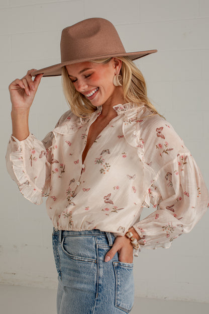 A woman wearing a beige long-sleeve blouse with a delicate pink and green floral print, styled with a brown wide-brim hat and blue jeans against a white brick wall.