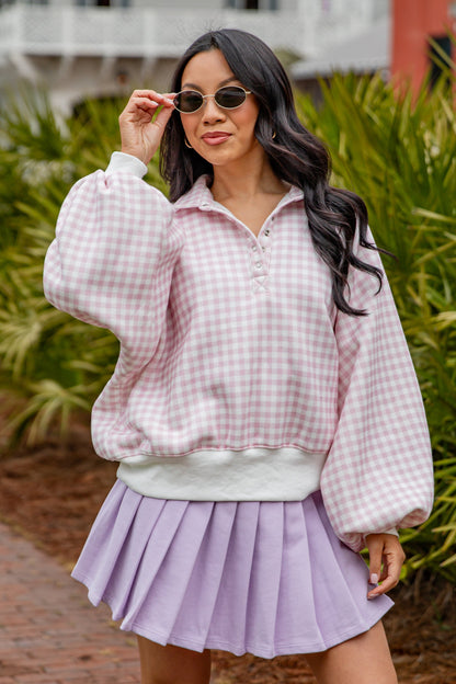 Woman wearing a checkered pink and white top and purple pleated skirt outdoors.