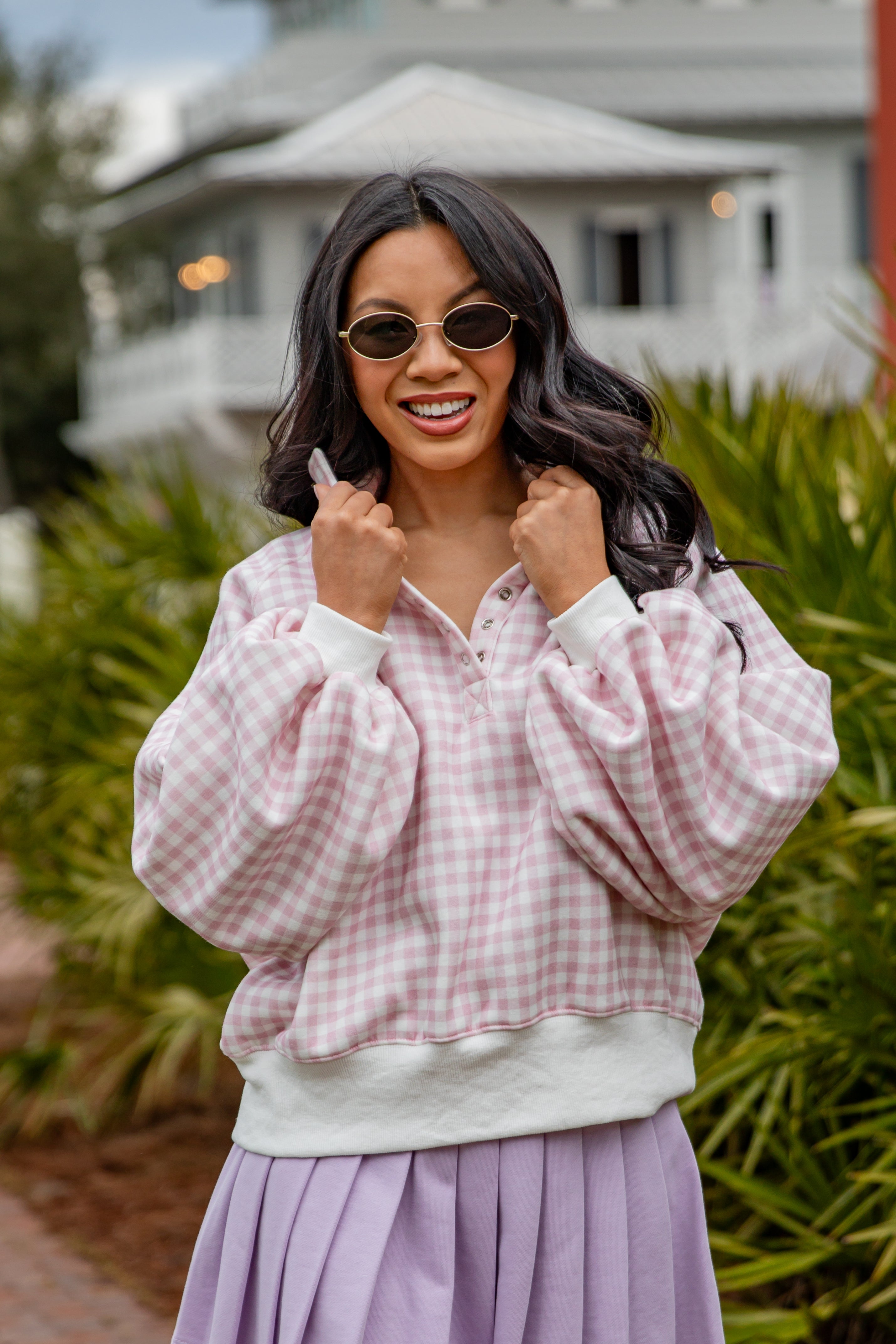 Woman wearing a pink checkered shirt over a white sweater with a purple skirt, standing outdoors.
