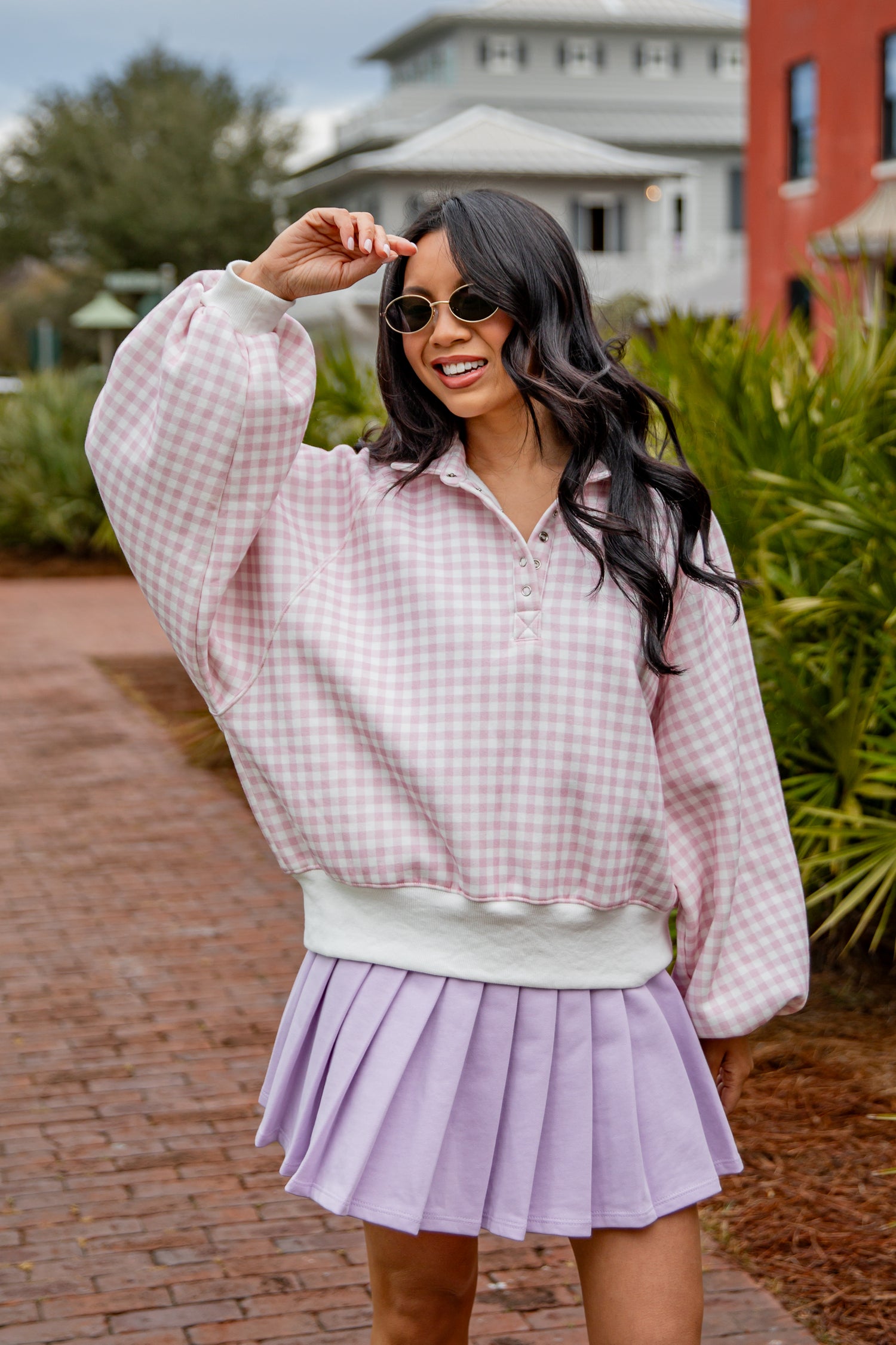Woman wearing a pink checkered blouse and purple pleated skirt outdoors.