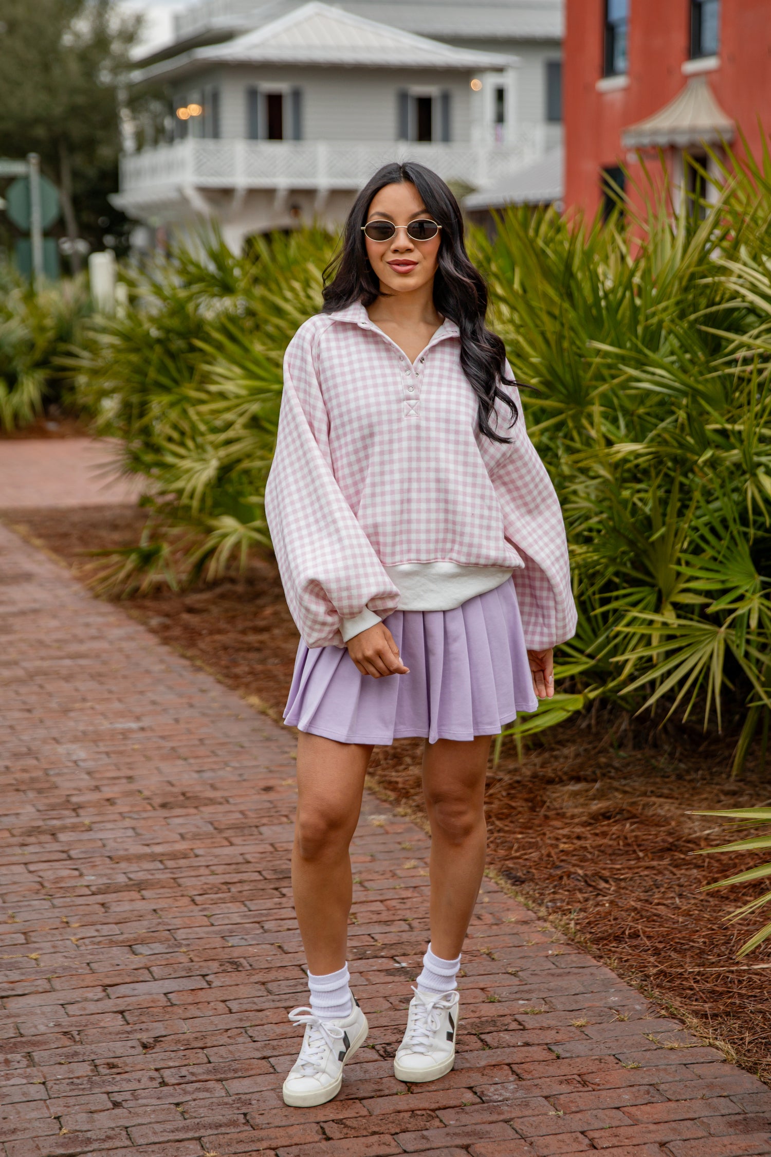Woman in a plaid shirt and purple skirt standing on a brick path with greenery and buildings in the background.