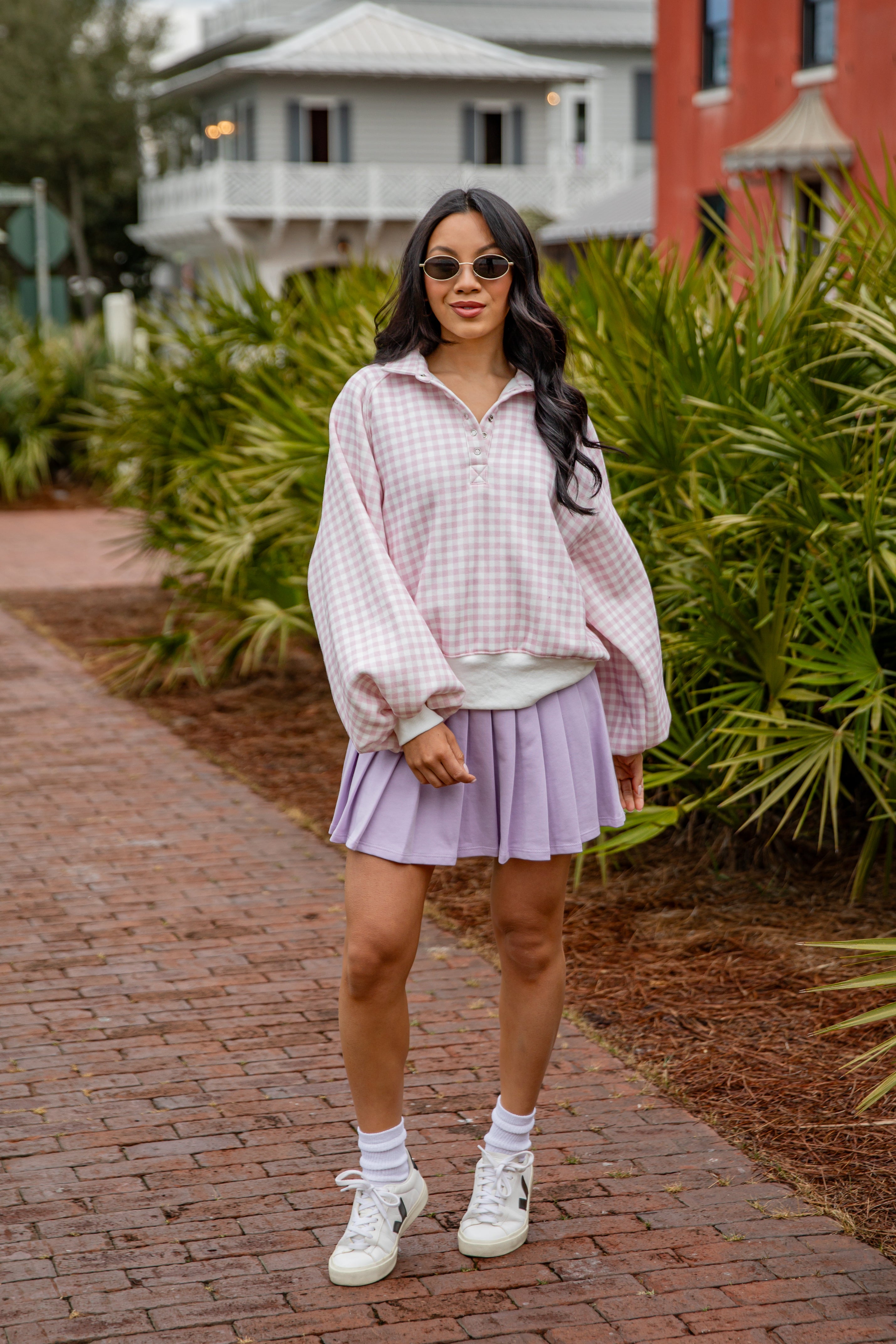 Woman in a plaid shirt and purple skirt standing on a brick path with greenery and buildings in the background.