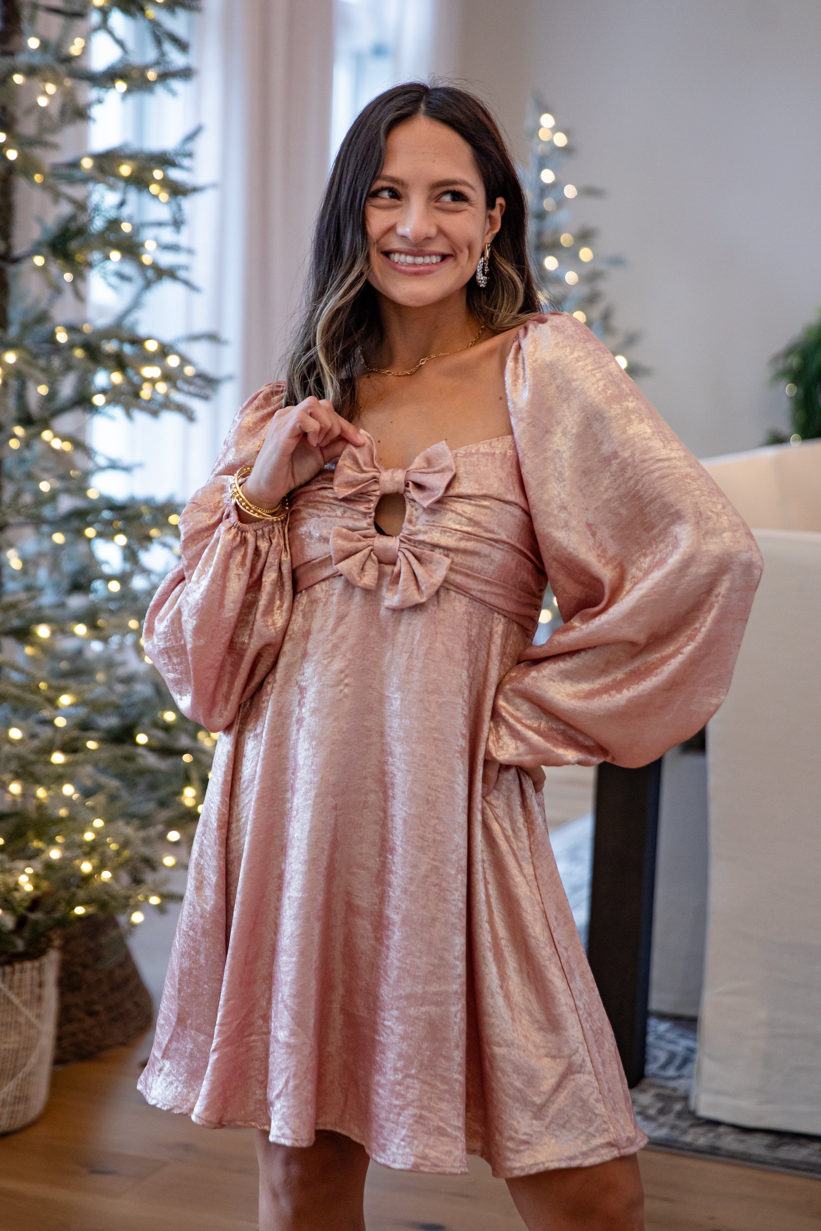 Woman wearing a pink dress with long sleeves and bow details, standing in a room with Christmas trees in the background.