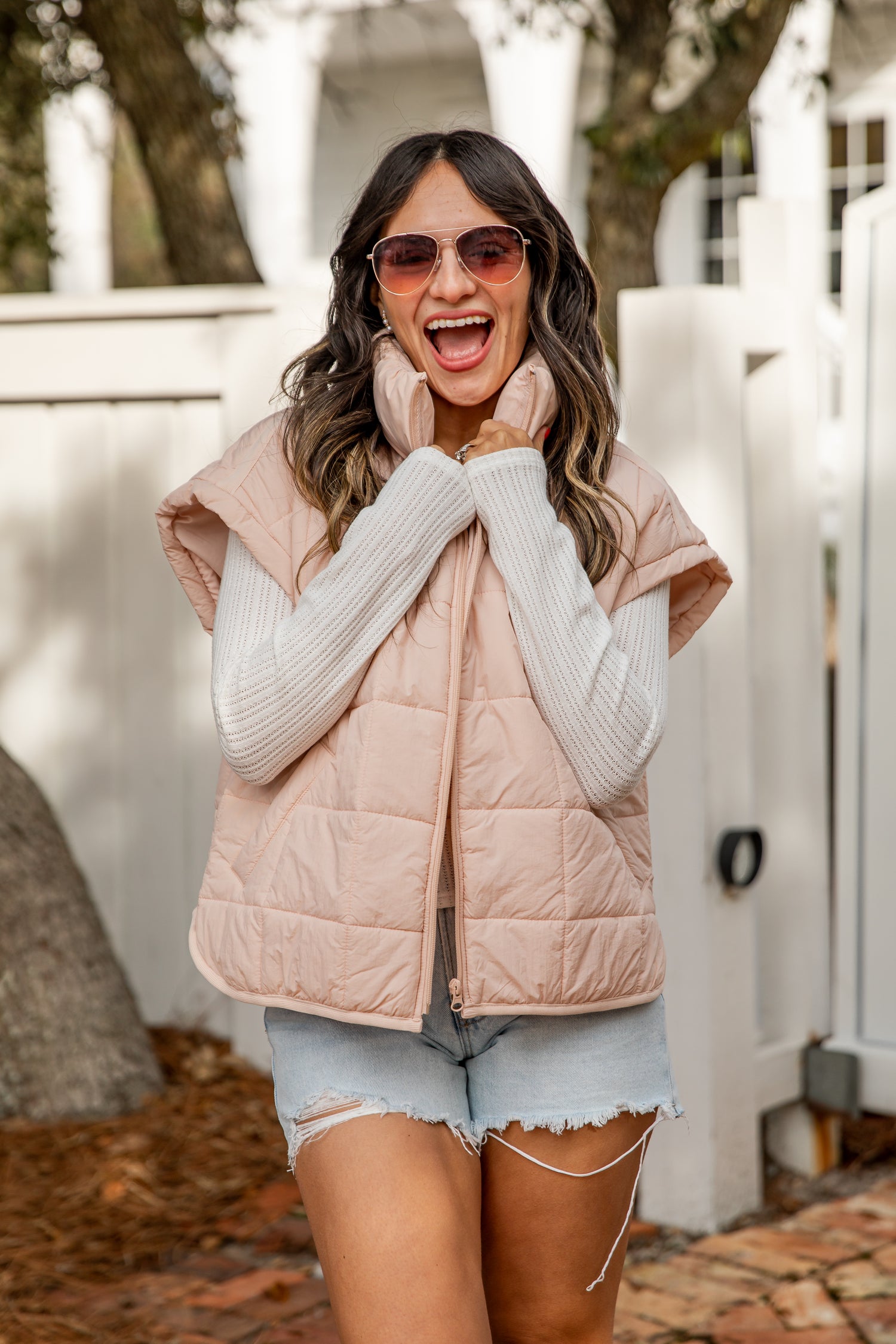 Woman wearing a light pink puffer vest over a white sweater with denim shorts, standing outdoors.