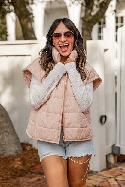 Woman wearing a light pink puffer vest over a white sweater with denim shorts, standing outdoors.