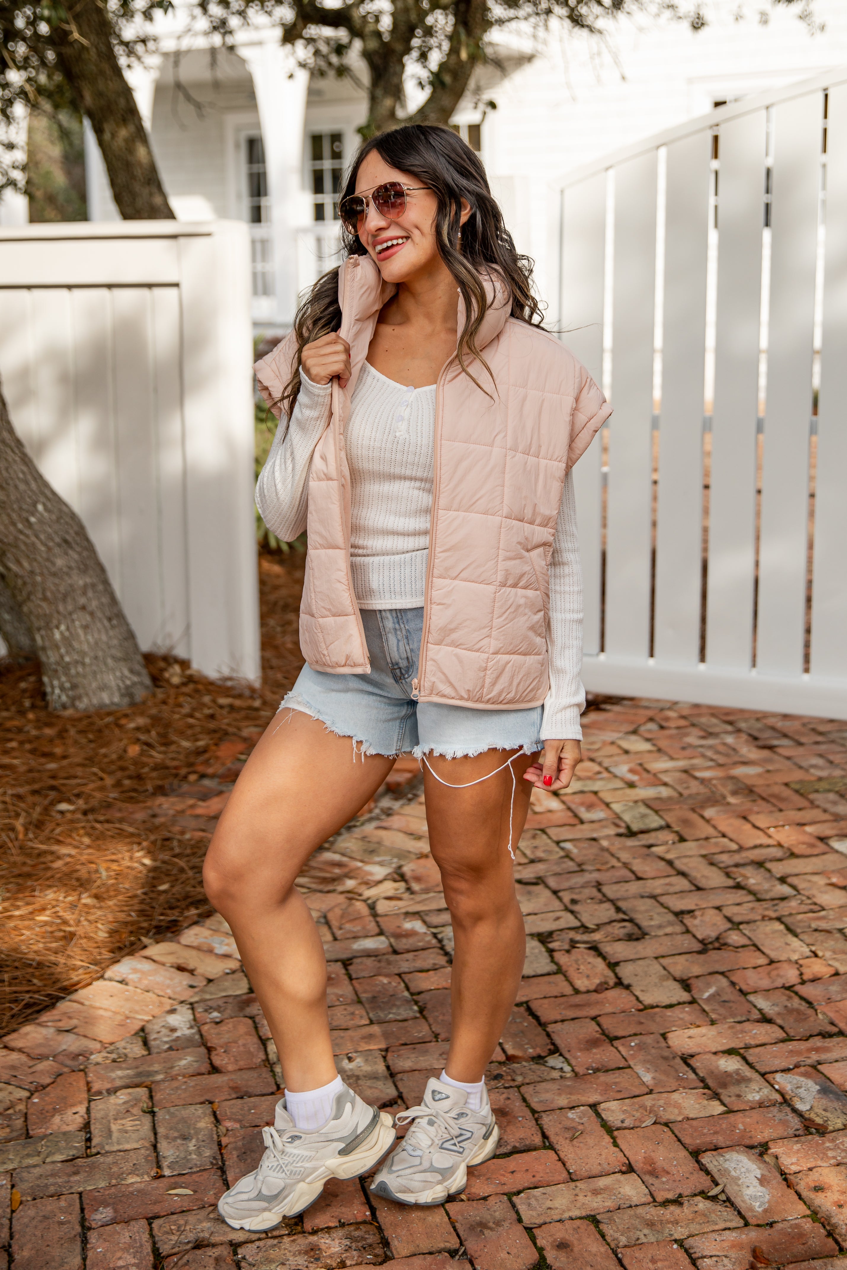 Woman wearing a light pink puffer vest, white sneakers, and denim shorts on a brick path.
