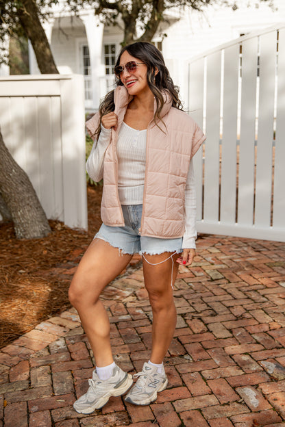Woman wearing a light pink puffer vest, white sneakers, and denim shorts on a brick path.