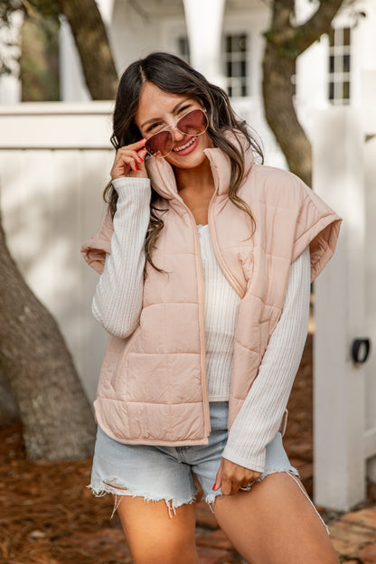 Woman wearing a light pink puffer vest over a white long-sleeve shirt and denim shorts, standing outdoors.
