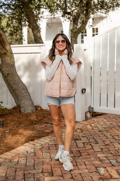 Woman wearing a pink puffer vest and denim shorts standing on a brick path.