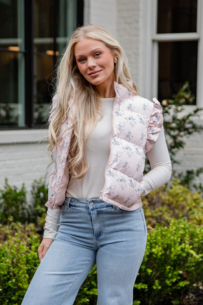 Woman wearing a floral puffer vest over a white shirt and blue jeans, standing outdoors.