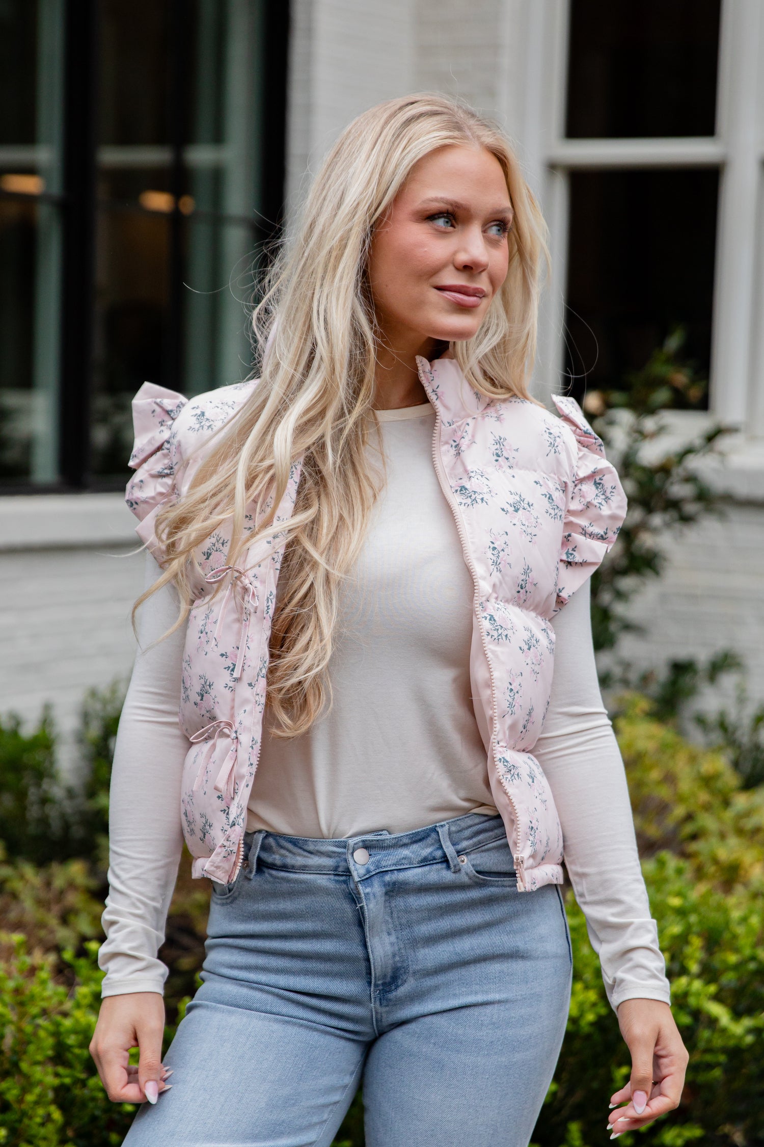 Woman wearing a light pink floral puffer vest over a white long-sleeve shirt and blue jeans, standing outdoors.