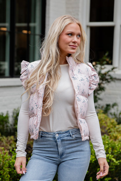 Woman wearing a light pink floral puffer vest over a white long-sleeve shirt and blue jeans, standing outdoors.