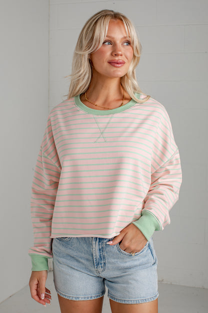 A woman wearing a pastel pink and mint green striped long-sleeve crewneck sweatshirt with solid mint green ribbed collar, cuffs, and hem, paired with light-wash denim shorts.