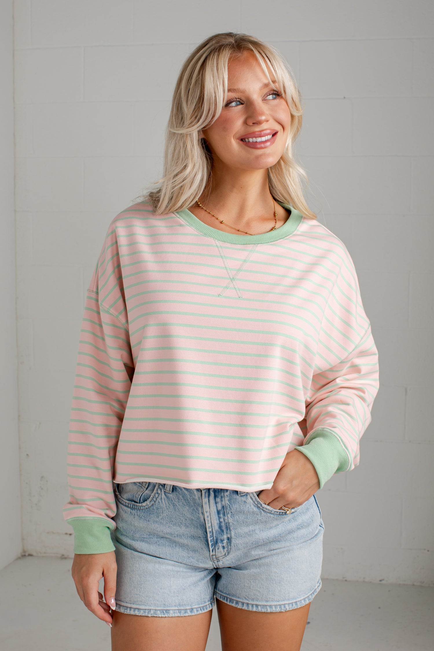 A woman wearing a pastel pink and white striped long-sleeve crewneck sweatshirt with sage green trim, paired with light-wash denim shorts, standing against a light gray brick wall.