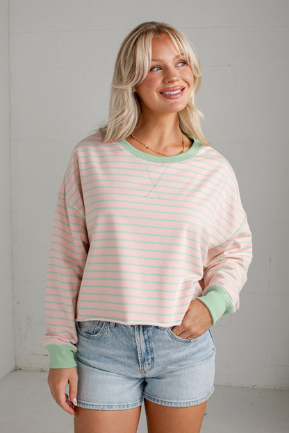 A woman wearing a pastel pink and white striped long-sleeve crewneck sweatshirt with sage green trim, paired with light-wash denim shorts, standing against a light gray brick wall.