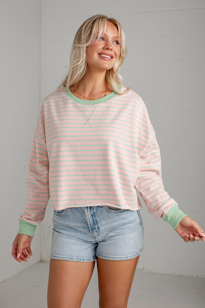 A woman wearing a light pink and white striped long-sleeve crewneck sweatshirt with sage green trim, paired with light-wash denim shorts, standing against a white paneled wall.