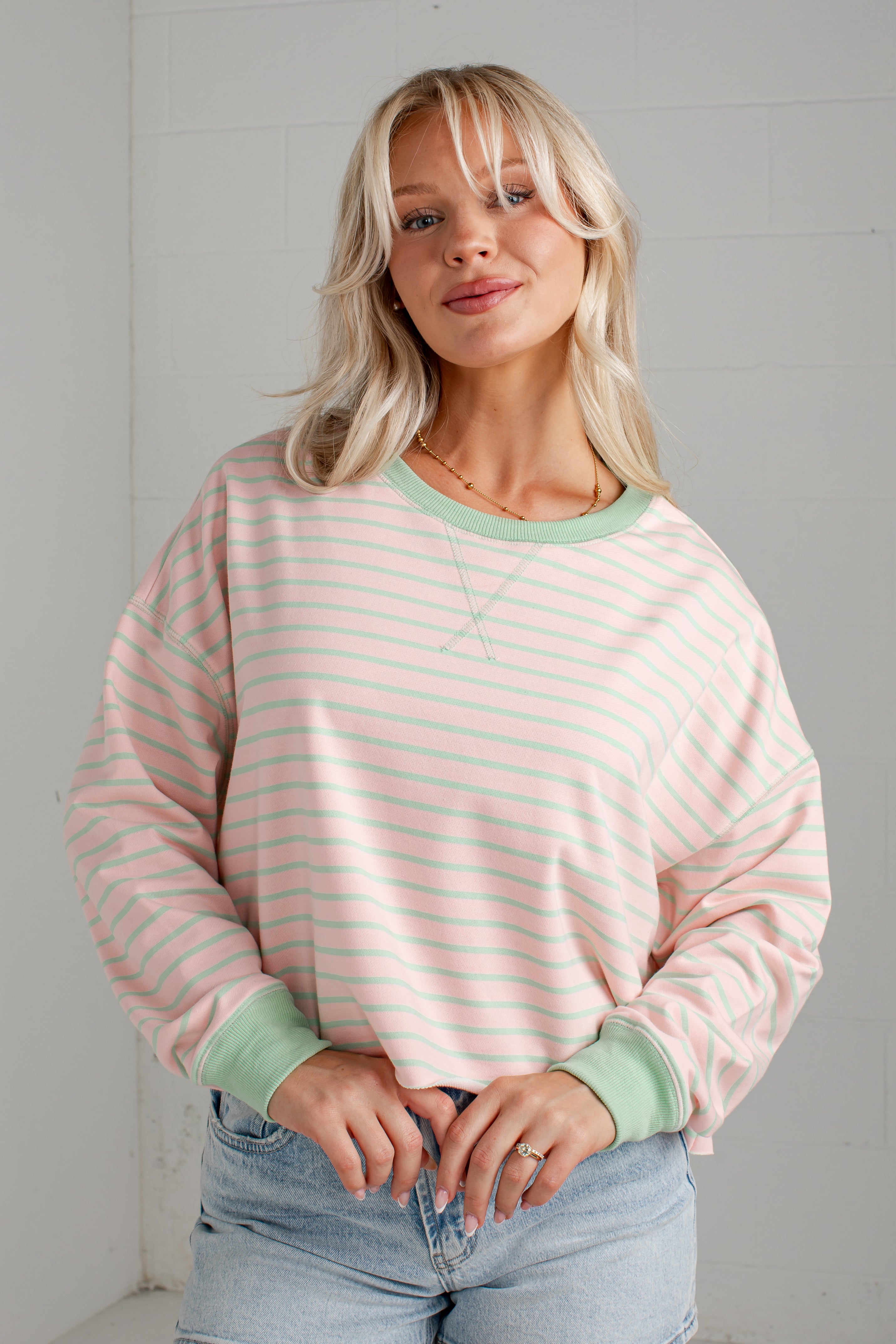 A blonde woman wearing a pastel pink and mint green striped long-sleeve sweatshirt with solid mint green ribbed collar, cuffs, and hem, paired with light-wash blue jeans.