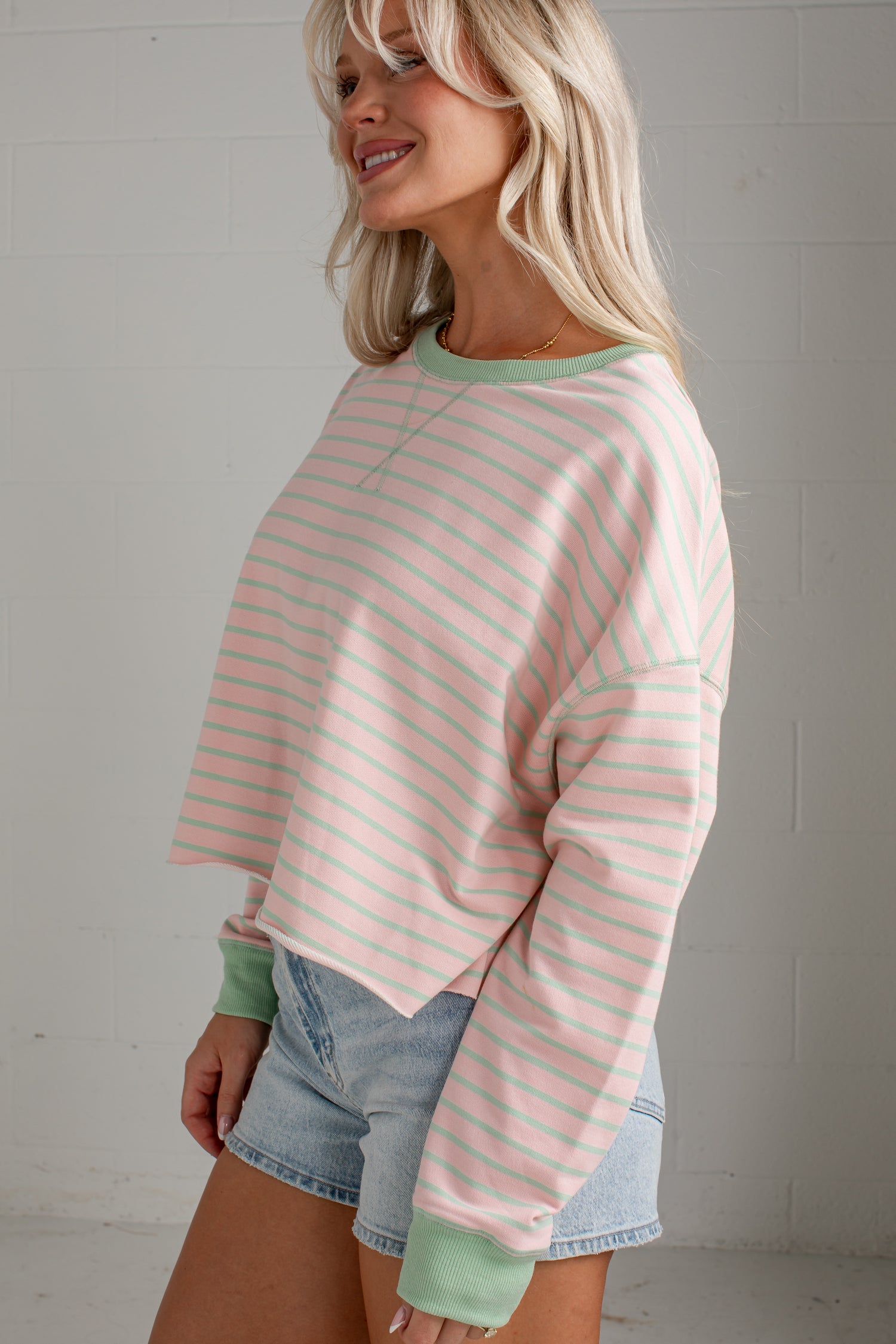 A woman wearing a pastel pink and mint green striped long-sleeve crop sweatshirt with solid green ribbed cuffs and collar, paired with denim shorts, standing against a white brick wall.