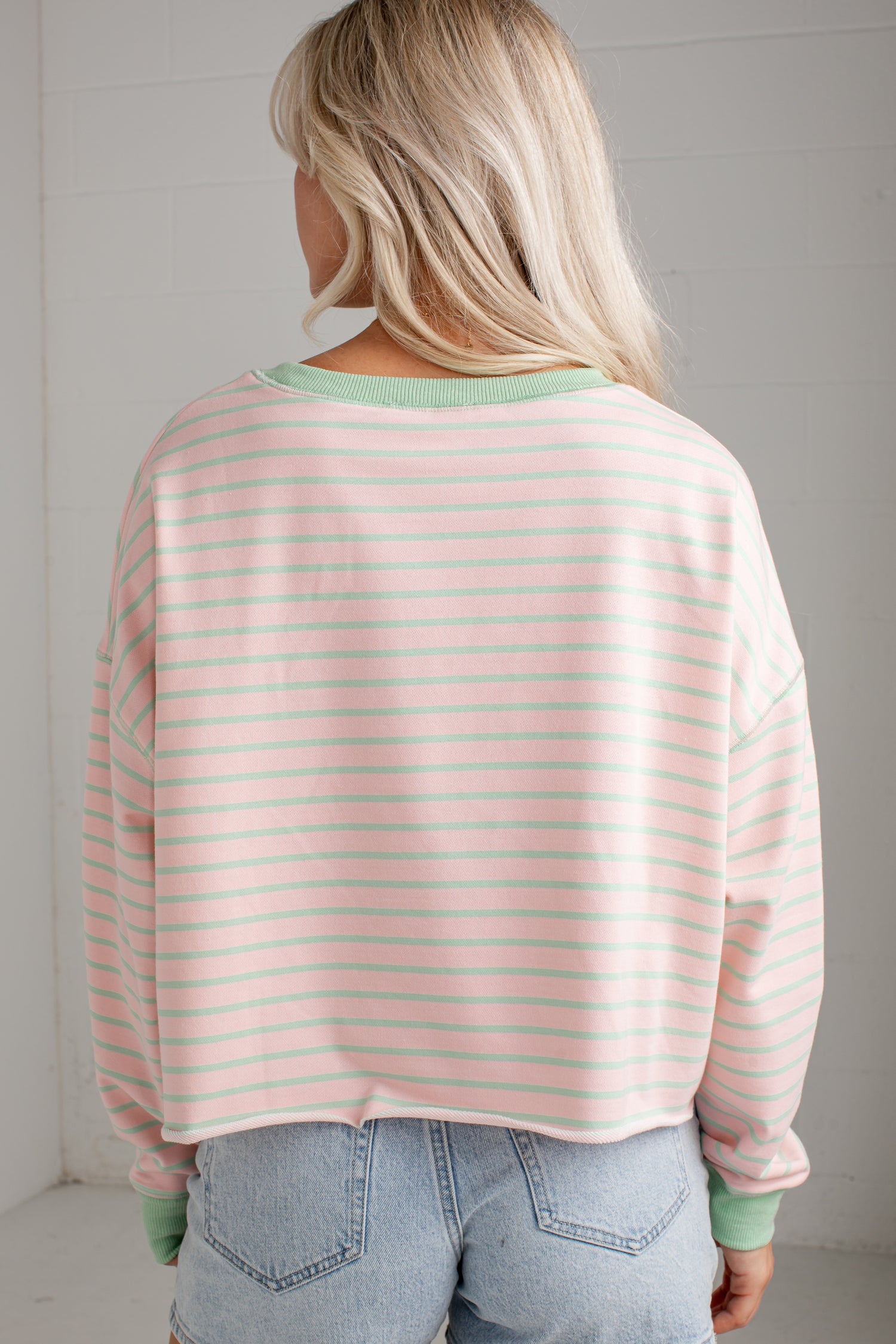 A woman viewed from behind wearing a light pink and white striped long-sleeve crop top with sage green ribbed trim, paired with light blue jeans.