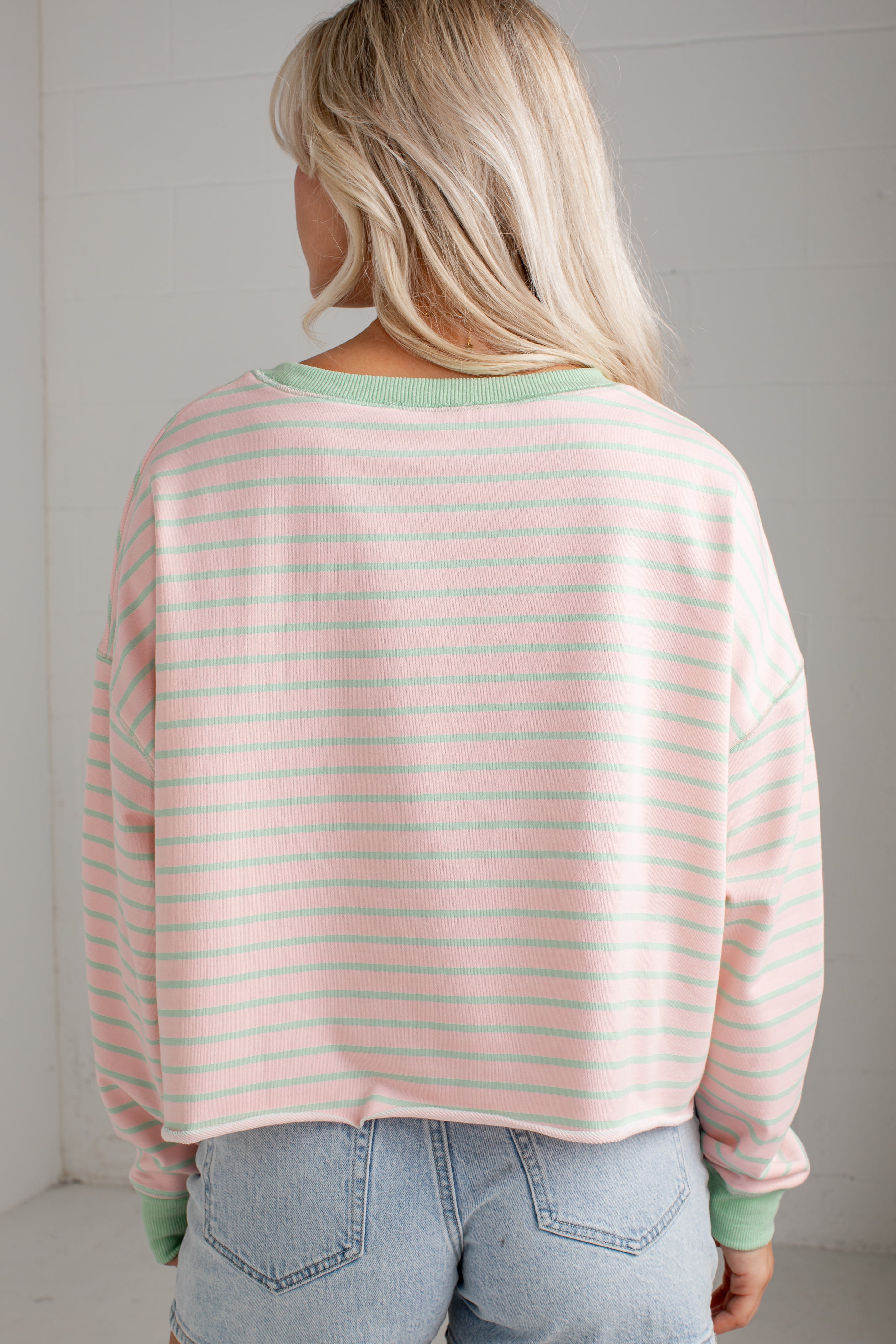A woman viewed from behind wearing a light pink and white striped long-sleeve crop top with sage green ribbed trim, paired with light blue jeans.