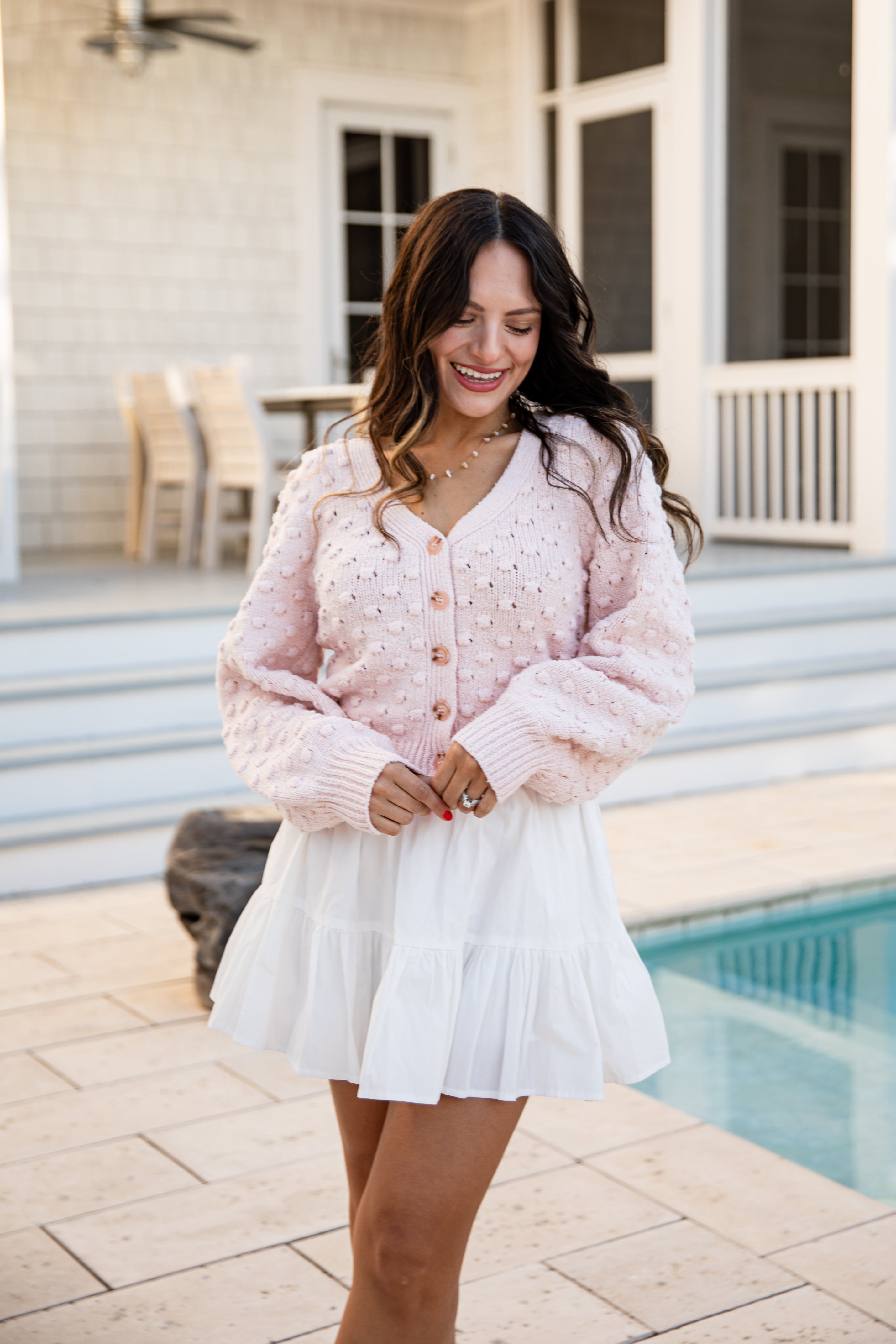Woman wearing a pink cardigan and white skirt standing by a pool.